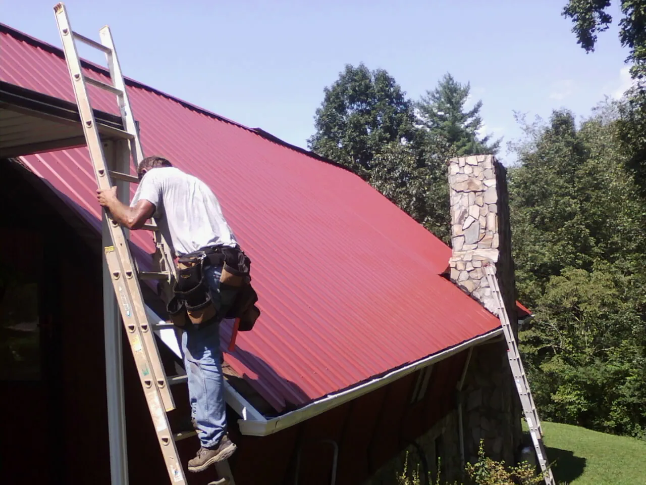 Roofer on a ladder beside a red metal roof and stone chimney — Soffit & Fascia Repair by Roofing Brothers in Barnwell